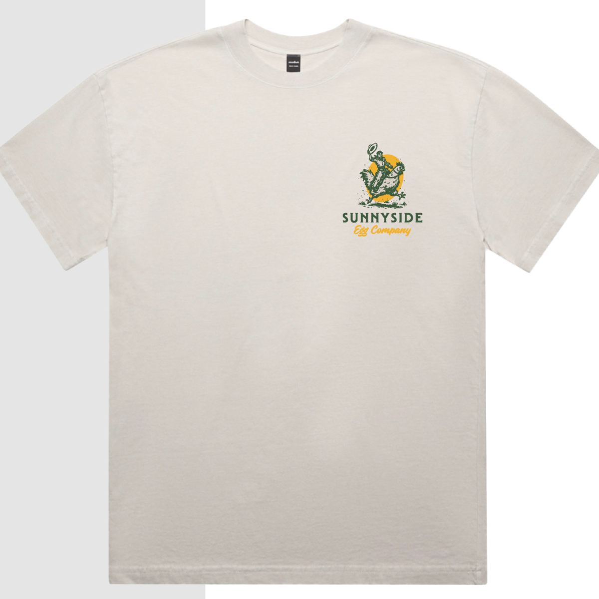 An off-white crewneck t-shirt featuring a green and yellow graphic of a cowboy riding a chicken with the text 'SUNNYSIDE Egg Company' printed below it.